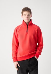 Crvena zip-up dukserica s visokim ovratnikom i raglan rukavima, s rebrastim rubom i manžetama. Mekana, glatka tekstura. Nositi s crnim hlačama.
