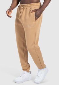 Beige Sweatpants mit elastischem Bund und seitlichen Taschen. Mit gestickten Logodetails. Getragen mit weißen Sneakers.