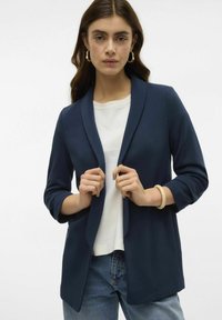 Blazer bleu marine avec col châle, en tissu texturé, porté sur un top blanc, associé à un jean bleu et un bracelet beige.