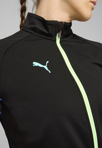 Zwarte sportjack met een hoge kraag, voorzien van een mintgroene rits en het Puma-logo. De stof is glad met contrasterende texturen aan de zijkanten.