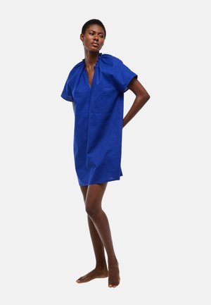 Femme debout pieds nus portant une robe bleue ample à manches courtes, longueur genou, avec un col froncé sur un fond uni.