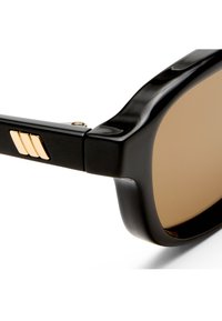 Κοντινό πλάνο μαύρων γυαλιών ηλίου με ορθογώνιους καφέ φακούς και χρυσή λεπτομέρεια με τριπλή ράβδο στο πλαϊνό μπράτσο.