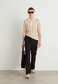 Μπεζ polo πουκάμισο με άσπρο γιακά, μαύρο παντελόνι και ανοιχτόχρωμα αθλητικά παπούτσια. Το μοντέλο κρατά μια μαύρη τσάντα και φορά γυαλιά ηλίου. Ουδέτερο φόντο.