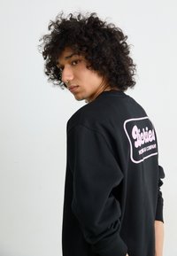 Felpa nera con maniche lunghe, caratterizzata da un grande logo rosa sul retro, realizzata in tessuto morbido, con una vestibilità rilassata e polsini a costine.