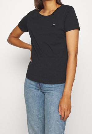 Kvinde iført en sort t-shirt med korte ærmer og et lille logo på brystet samt lyseblå jeans, der står med den ene hånd på hoften.