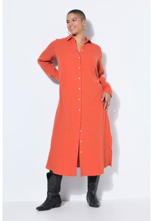 Smilende kvinde iført lang orange skjortekjole med knapper og sorte støvler til midt på læggen, stående med den ene hånd på hoften.