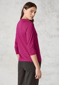Fuchsia Langarmshirt mit geometrischem Muster, Rundhalsausschnitt und 3/4-Ärmeln, kombiniert mit dunkelgrauen Hosen.