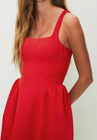 Rode mouwloze jurk met een rechte halslijn, nauwsluitend lijfje en een geplooide rok met zijzakken. Zachte textuur, gladde stof.