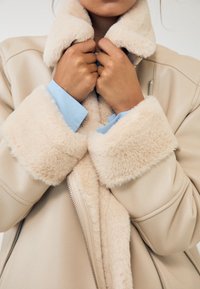 Béžová bunda z umělé kůže s huňatým béžovým límcem a manžetami, vybavená předním zipem a světle modrou košilí pod ní, ukazující kontrast textur.