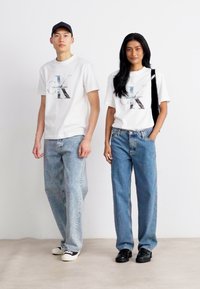 Dva jedinci mají na sobě shodné bílé trička s logem Calvin Klein. Kombinují je s volnými světle modrými džíny a černými botami.