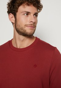 Roter Baumwollpullover mit rundem Ausschnitt und gerippten Bündchen. Verfügt über ein kleines gesticktes Logo auf der Brust. Glatte Textur.