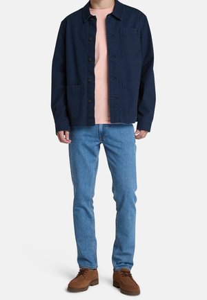 Veste bleu marine avec fermeture boutonnée et deux poches avant, portée sur un t-shirt rose clair, associée à un jean bleu clair et des chaussures marron.