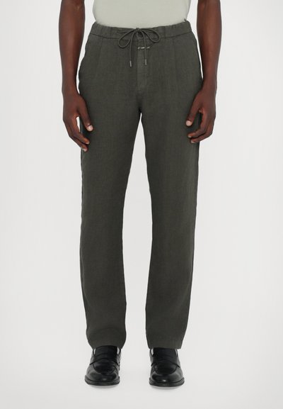Pantalons en lin vert foncé avec taille à cordon, devant plissé et coupe décontractée. Associés à des chaussures habillées noires.
