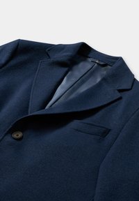 Granatowa marynarka z wełny z wyciętym klapem, zapinana na jeden guzik, z kieszenią na piersi. Teksturowana tkanina nadaje głębi wizualnej.