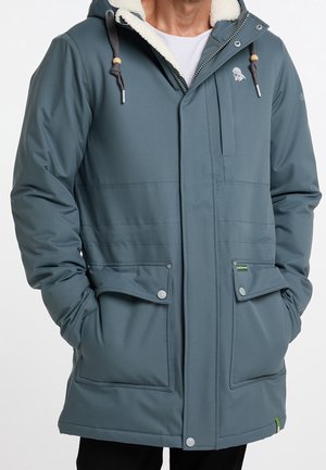 Homme portant un manteau d’hiver bleu à capuche avec doublure polaire, fermeture éclair à l’avant, poches à rabat boutonnées et capuche à cordon, mains dans les poches.