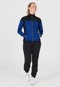 Trackjakke i blå og sort stof med lynlås-lukning, parret med sorte bukser, der har elastiske manchetter og hvide sportssko.