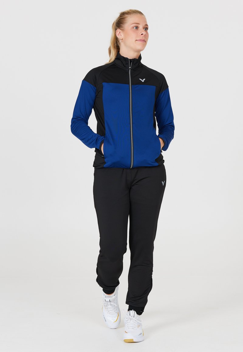 Trackjakke i blå og sort stof med lynlås-lukning, parret med sorte bukser, der har elastiske manchetter og hvide sportssko.