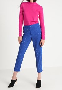 Kirkkaan vaaleanpunainen neulepaita korkealla kaula-aukolla, yhdistetty sinisiin housuihin, joissa on pistekuvio, ja mustiin korkokenkiin.