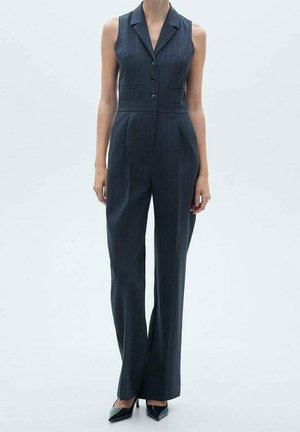 Femme portant une combinaison sans manches à fines rayures sombres avec des boutons, pantalon à jambes larges et talons hauts noirs pointus.
