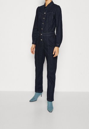 Kobieta ubrana w ciemnoniebieski kombinezon dżinsowy z długimi rękawami, zapinany na guziki, oraz jasnoniebieskie botki na obcasie, na jednolitym tle.