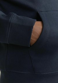 Tengerészeti pulóver ribbed mandzsettákkal és kenguruzsebbekkel, sima textúrával és egy kéz közelképe, ami a zsebben pihen.