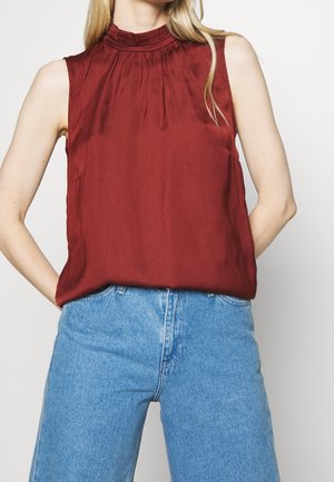 Vrouw draagt een mouwloze bordeauxrode top met een hoge hals, ingestopt in een lichtblauwe, hooggetailleerde spijkerbroek, staat met handen achter de rug.