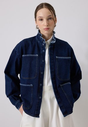 Tmavomodrá rifľová bunda s vysokým golierom, dvoma veľkými prednými vreckami, kontrastným bielym prešívaním a zapínaním na gombíky vpredu.