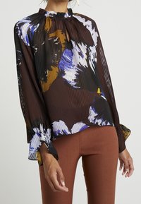 Durchsichtige braune Bluse mit bunten Blumenmustern, plissierter Textur, Rüschenausschnitt und Glockenärmel, kombiniert mit taillierten braunen Hosen.