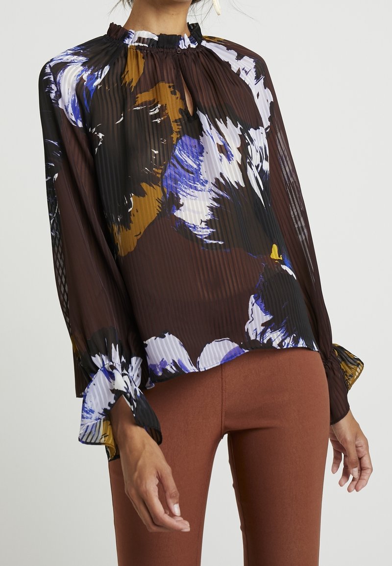 Durchsichtige braune Bluse mit bunten Blumenmustern, plissierter Textur, Rüschenausschnitt und Glockenärmel, kombiniert mit taillierten braunen Hosen.