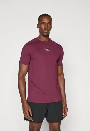 Bordeaux farvet trænings T-shirt med korte ærmer, rund halsudskæring og diskret logo på brystet. Glat tekstur, tætsiddende design. Sammen med sorte shorts.
