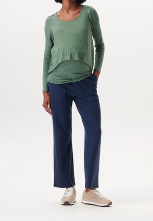 Vrouw draagt een groene, lange mouw, gelaagde zwangerschaps top, donkerblauwe broek en beige sneakers, met één hand in de zak, staand op een witte achtergrond.