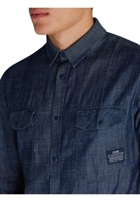 Granatowa koszula dżinsowa z kołnierzykiem na guziki, dwoma kieszeniami na piersiach oraz markową metką. Tkanina ma teksturowany wygląd.