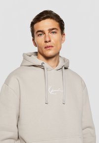 Haut à capuche beige clair avec cordon de serrage et poche avant. Présente un logo brodé blanc sur la poitrine. Texture de tissu douce.