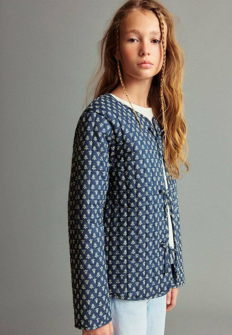 Mladostnik z dolgimi pričeskami, ki nosi modro vzorčasto prešito jakno in svetlo modre kavbojke, stoji ob enobarvni sivi podlagi.