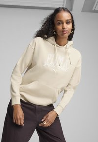 Grå hoodie med en frontlomme, der har et hvidt "PUMA sports club" grafik. Blødt stof og en afslappet pasform. Mørke bukser bæres under.