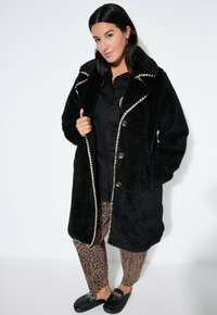Schwarzer, strukturierter Kunstfellmantel mit großem Kragen, cremefarbenem Besatz und Knöpfen. Kombiniert mit Hosen im Leopardenmuster und schwarzen Slippers.