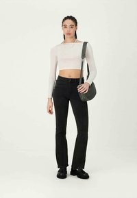 Mulher com cabelo trançado, vestindo um top cropped de manga longa branco, jeans preto de cintura alta, sapatos pretos e a carregar uma mala de ombro cinza-escuro.