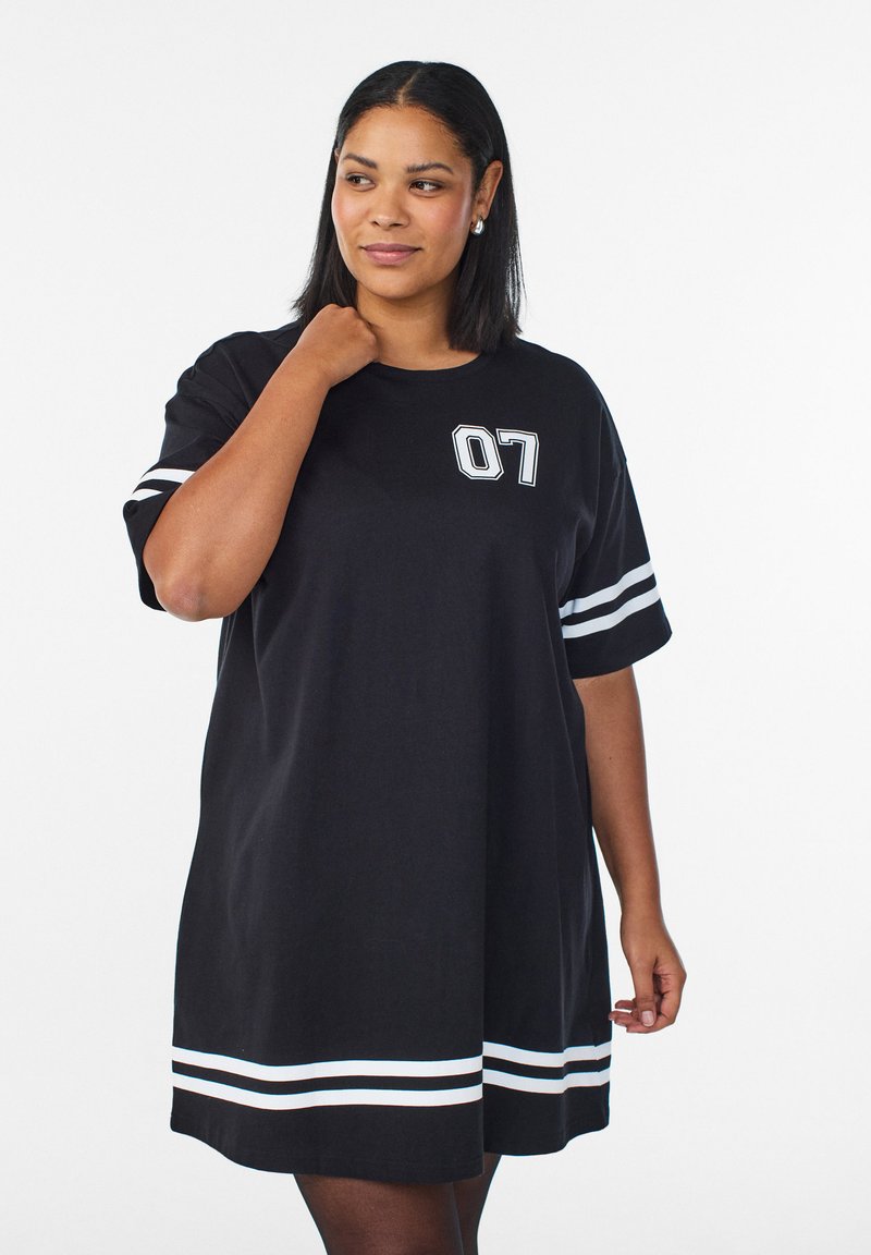 Femme portant une robe T-shirt noire oversize avec des rayures blanches sur les manches et l'ourlet, et le numéro 07 imprimé sur la poitrine.