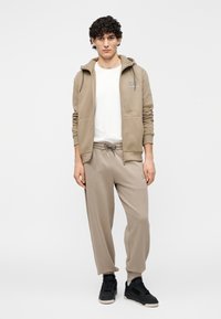 Beige hoodie met rits en zakken, gecombineerd met bijpassende beige joggingbroek met trekkoord. Witte basic t-shirt eronder. Zwarte sneakers.