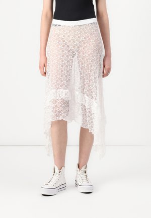 Jupe en dentelle blanche avec un ourlet asymétrique, présentant un design en forme de scallop et une finition texturée, associée à des baskets montantes blanches.