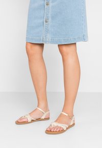 Beige litte sandaalit, joissa on punotut hihnat, avokärkinen muotoilu ja nilkkahihna. Yhdistetty vaaleansinisen farkkuhameen kanssa, jossa on napit.