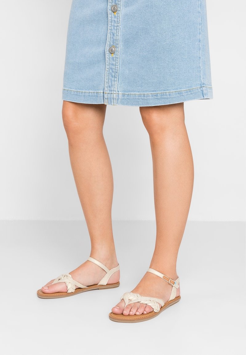 Beige litte sandaalit, joissa on punotut hihnat, avokärkinen muotoilu ja nilkkahihna. Yhdistetty vaaleansinisen farkkuhameen kanssa, jossa on napit.