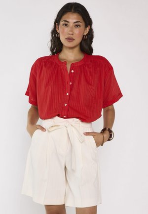 Femme portant un chemisier rouge à manches courtes boutonné et un short blanc taille haute avec une ceinture nouée, debout les mains dans les poches.