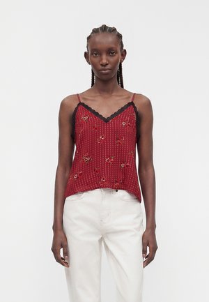 Femme aux cheveux tressés portant un haut sans manches rouge à fleurs avec une bordure en dentelle noire et un pantalon blanc taille haute, debout devant un fond uni.