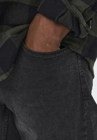 Jean noir en denim avec une poche avant, associé à une chemise à carreaux verte et noire. Le tissu présente une texture et un poids typiques des vêtements décontractés.