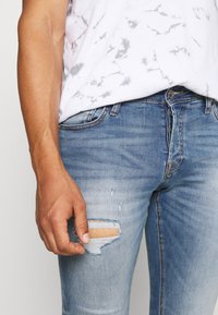 Blå denimjeans med slitna områden, inklusive ett stort hål vid knäet och subtil blekning. I kombination med en vit mönstrad skjorta.