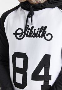 Felpa con cappuccio bianca e nera, caratterizzata da una texture a rete, con il logo "SikSilk" e un grande "84" in nero sulla parte anteriore.