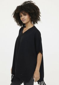 Schwarzer Strick-Poncho mit V-Ausschnitt und Fransenbesatz, ausgestattet mit kurzen Ärmeln und einer lockeren Passform. Minimalistisch in Stil und Konstruktion.