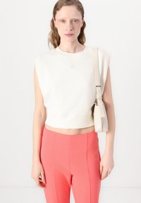 Kreminė trumpa sportinė palaidinė su storais rankovių iškirpimais ir DKNY logotipu; derinama su ryškiai koralinėmis aukštu liemeniu kelnėmis; modelis laiko mažą smėlio spalvos krepšį.