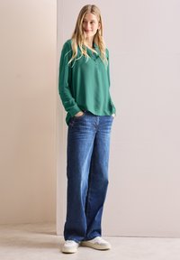 Camicia verde a maniche lunghe con colletto abbottonato, abbinata a jeans blu in denim morbidi e sneakers bianche su uno sfondo chiaro.